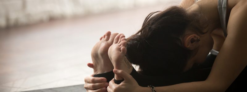 Young sporty yogi woman practicing yoga concept, sitting in paschimottanasana exercise, Seated forward bend pose, working out on black mat, studio floor background, legs and hands close up